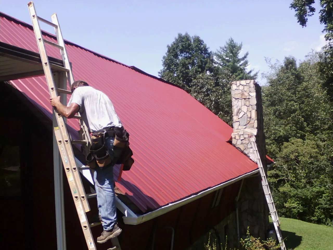 Roofer on a ladder beside a red metal roof and stone chimney — Soffit & Fascia Repair by Roofing Brothers in Mahwah